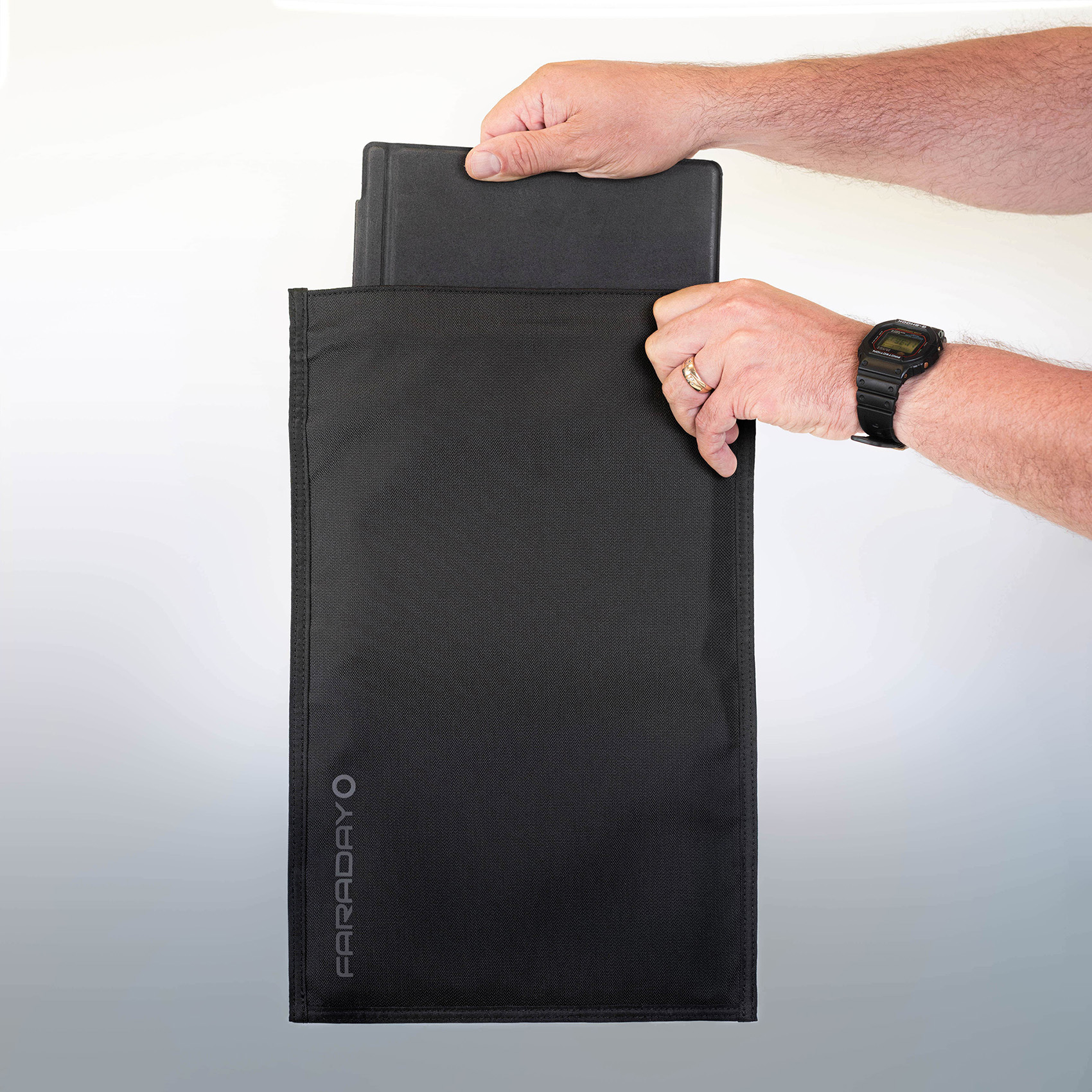 Homme tenant un étui noir en tissu rigide, glissant une tablette ou notebook à l’intérieur; logo Faraday sur le côté et bracelet noir au poignet.